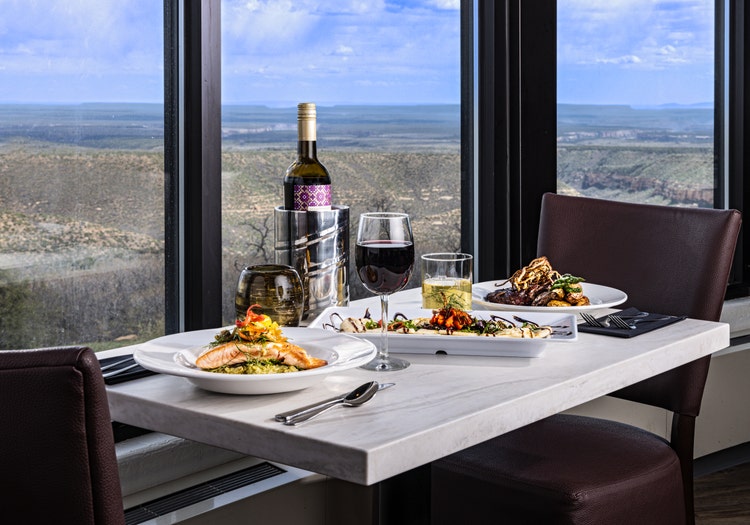 A modern restaurant table is set with gourmet dishes and drinks beside large windows overlooking a vast canyon landscape